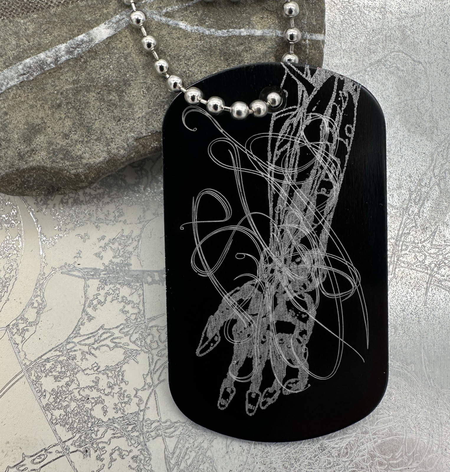 Black engraved dog tag with fine anatomical and looping linework.