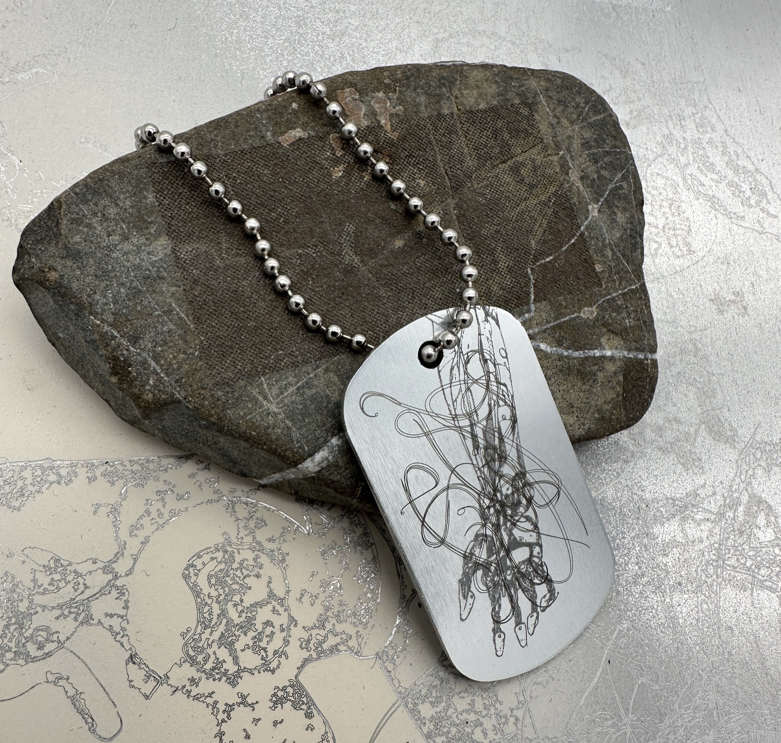 Silver-coloured engraved dog tag with anatomical and looping linework.