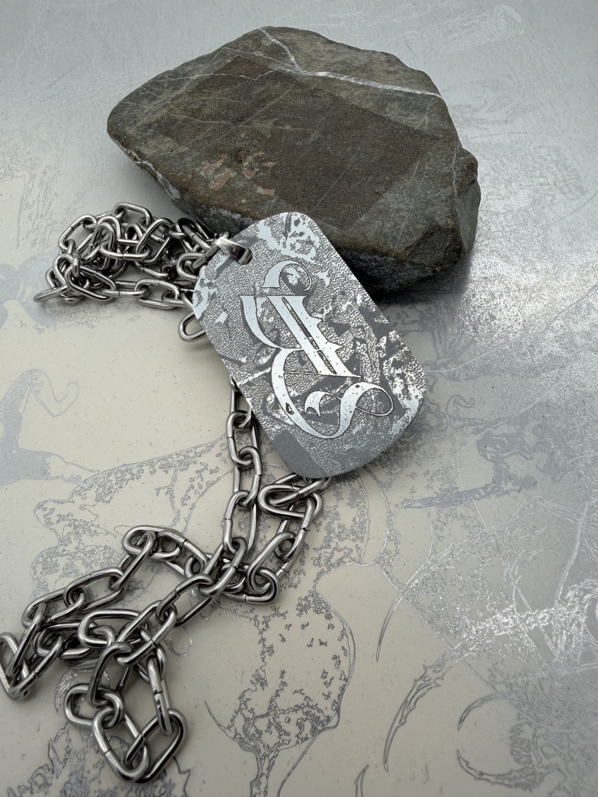 Silver-coloured chain with an engraved rectangular metal pendant bearing Basilisk lettering and textured imagery, resting beside a stone.