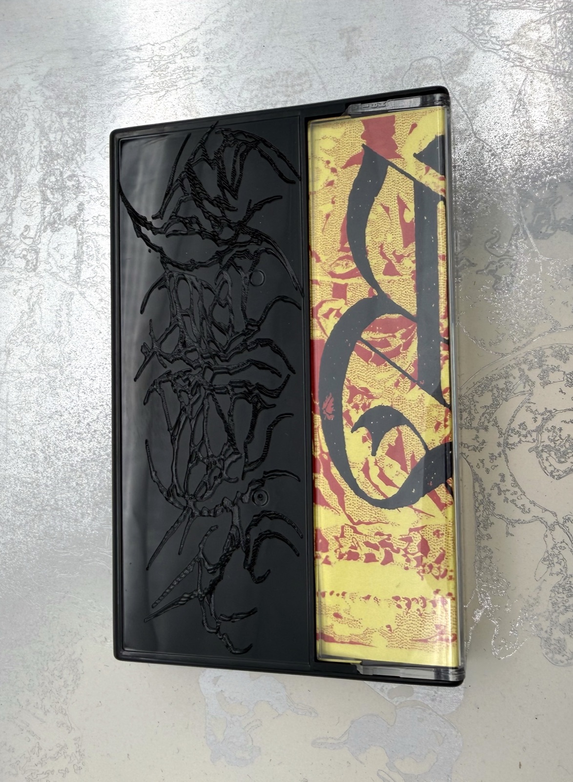 Black plastic cassette case with dark engraved branching lettering beside a yellow and red printed insert.
