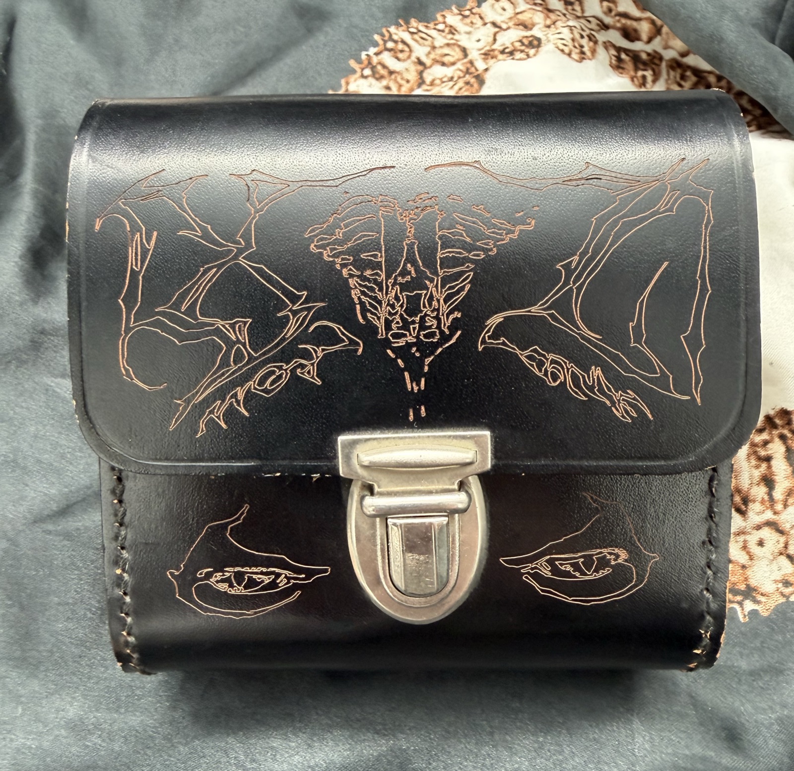 Front of a found black leather postal case with pale engraved linework, two eyes and an anatomical motif around a metal clasp.