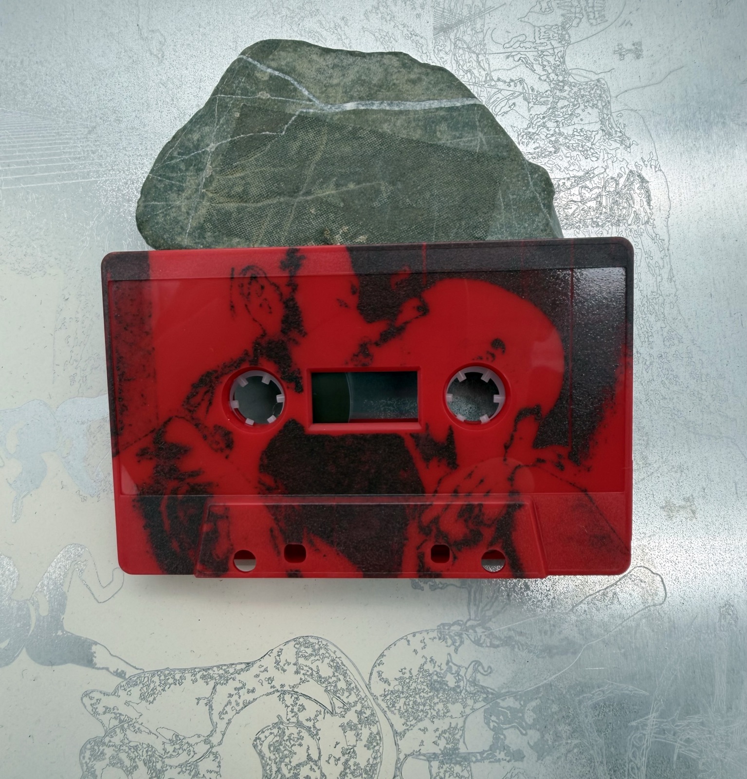 Red plastic audio cassette with dark photographic imagery extending across its shell.